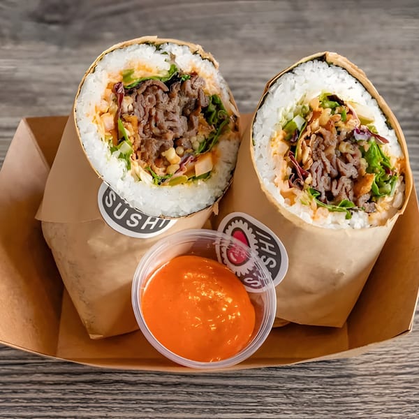 Two large sushi burritos with beef and vegetables, wrapped in paper, with a side of orange sauce.